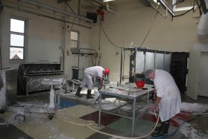 Two employees in a white coat or jumpsuit spray water and foam cleaner on the floor and various pieces of equipment in the Muscle Biology Laboratory.